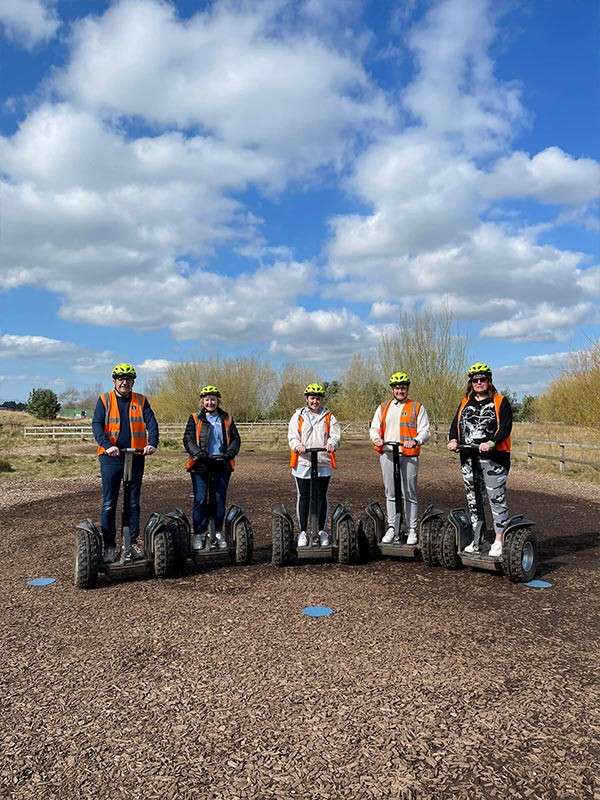 Five people on off-road Segways pose, hands on handlebars, on a woodchip track. They wear helmets and orange vests, with fencing, trees, and a partly cloudy sky.