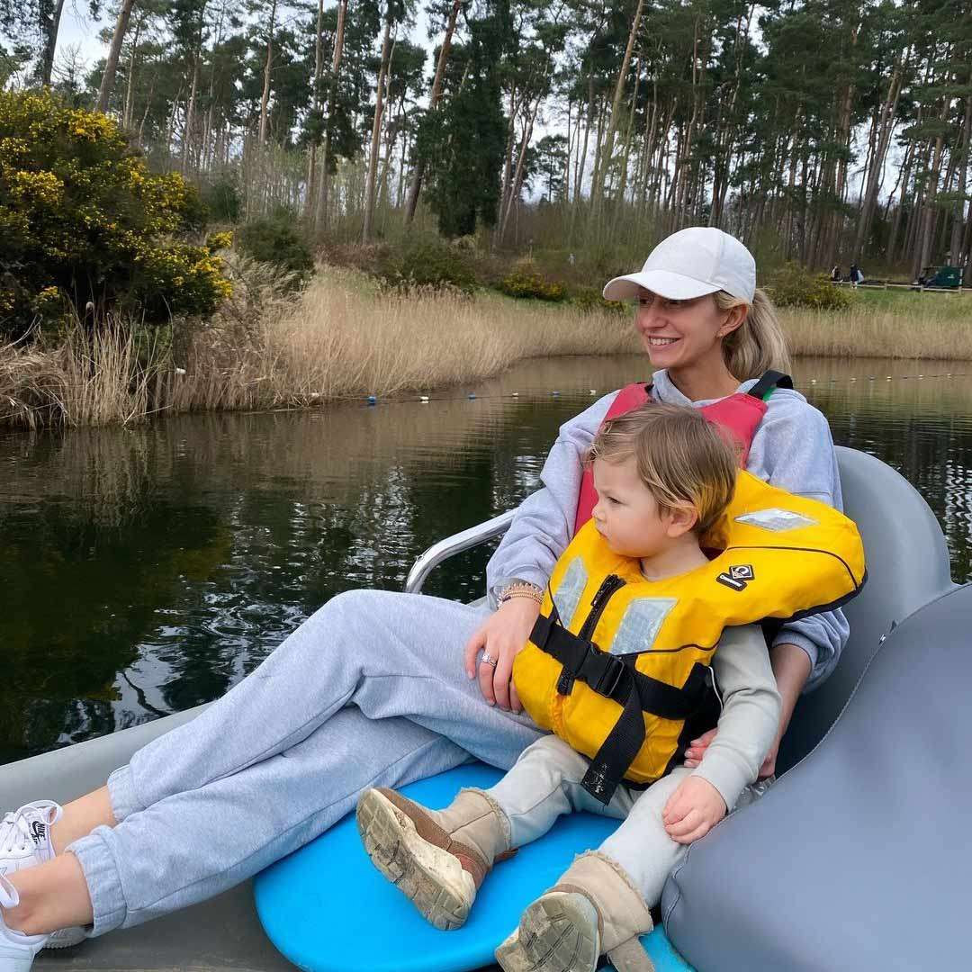 Child wearing yellow life jacket sits on an adult’s lap, both relaxing on a pedal boat on a calm lake, surrounded by reeds and tall pine trees under an overcast sky.