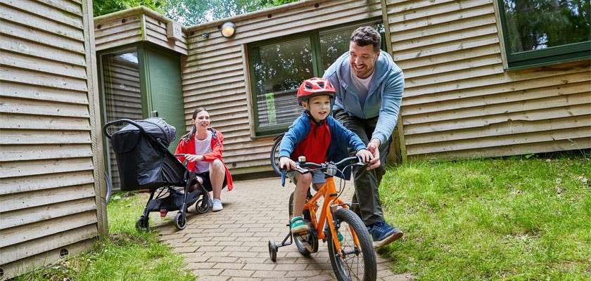 Child on an orange bicycle rides as a man steadies him; nearby, a woman with a stroller watches. Wooden cabins and a brick path lie amid grass and trees.