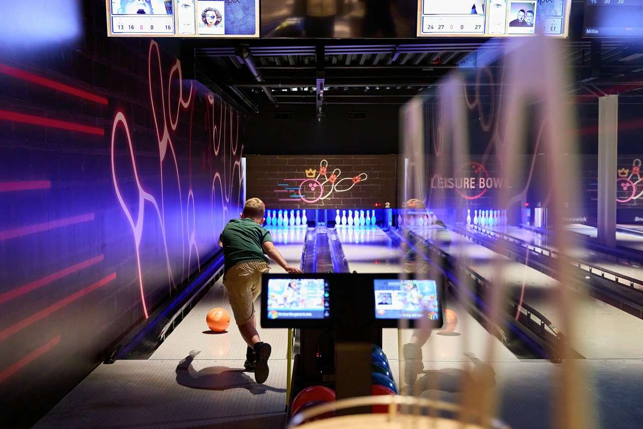 Bowler releases orange ball toward glowing pins; context: neon-lit bowling alley with digital screens and partitions, wall text "LEISURE BOWL", lane number "2", and brand "Brunswick".