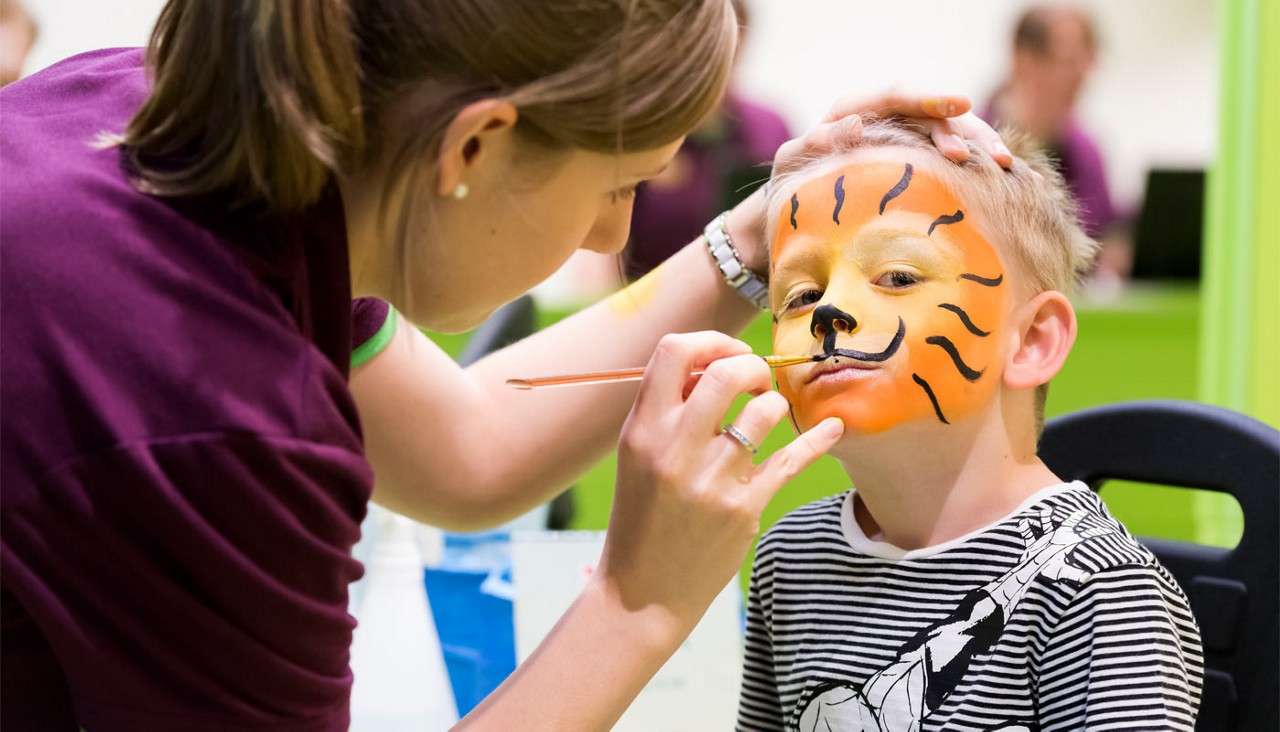 Child sits still while an artist paints a tiger face on him, brush at his lips; indoors with bright green walls and others blurred in the background.