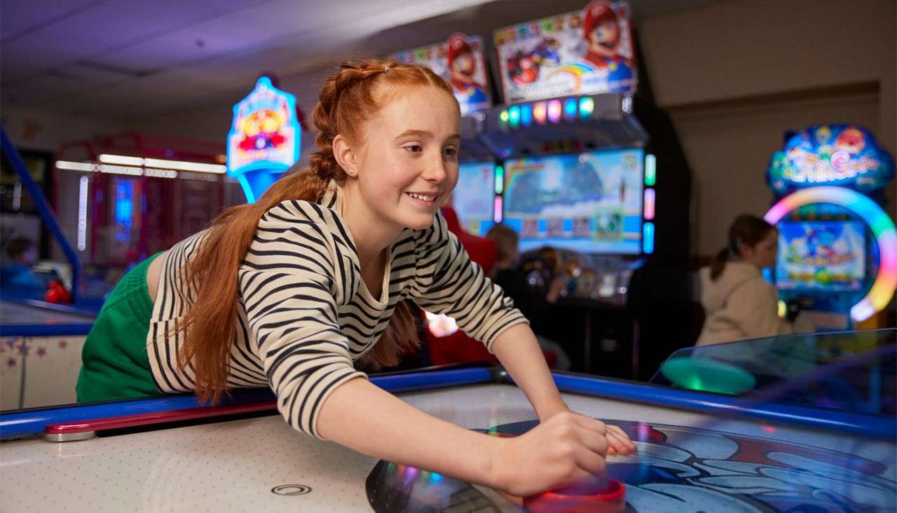 Child plays air hockey, pushing a red mallet; context: brightly lit arcade with racing-game cabinets and colorful lights in the background. Text: 10.