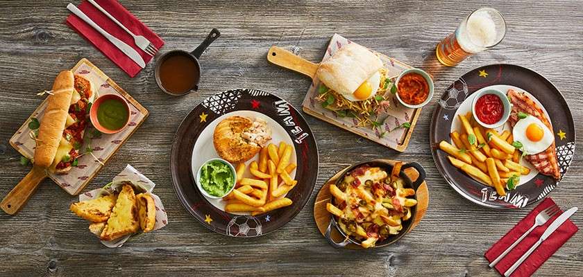 Assorted pub dishes are arranged on a wooden table: sandwiches, fries with dips, cheesy loaded fries, eggs, guacamole, and a pint of beer on soccer-themed plates. Text: TEAM!