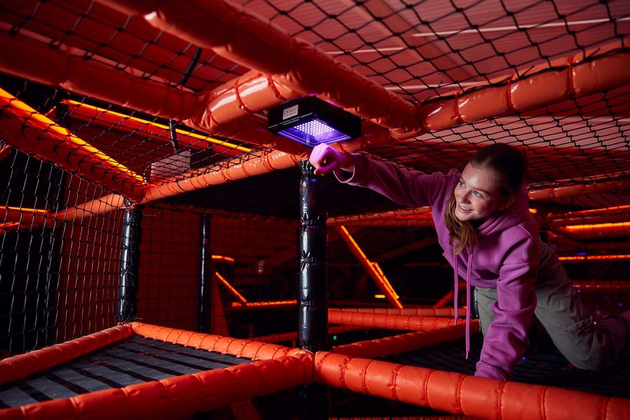 Child crawls on a netted platform, reaching to tap a small purple-lit target beneath an overhead beam, within an indoor climbing maze of orange-padded frames and dark, neon-lit surroundings.