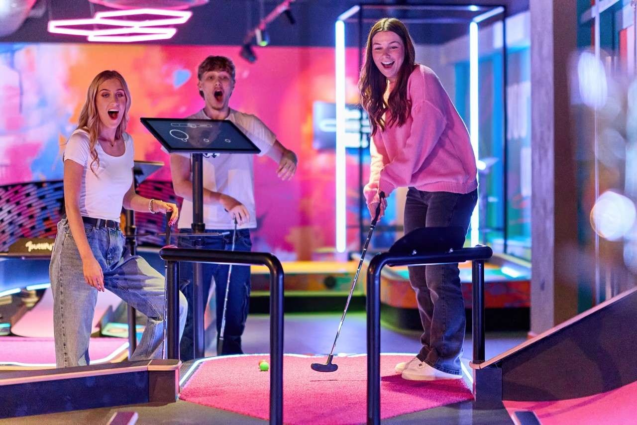 Young woman putts a ball on a pink mini-golf hole while two friends watch, reacting excitedly; neon lighting, touchscreen score kiosk, and rails define an indoor arcade-style mini-golf course.