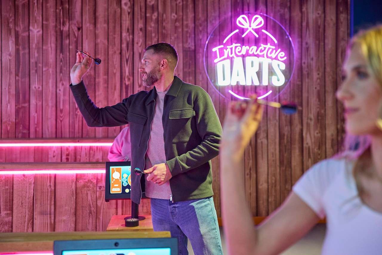 Man aims a dart, preparing to throw; another person does the same in foreground; neon sign reads "Interactive DARTS" on wooden wall; screens and strip lights complete the arcade setting.