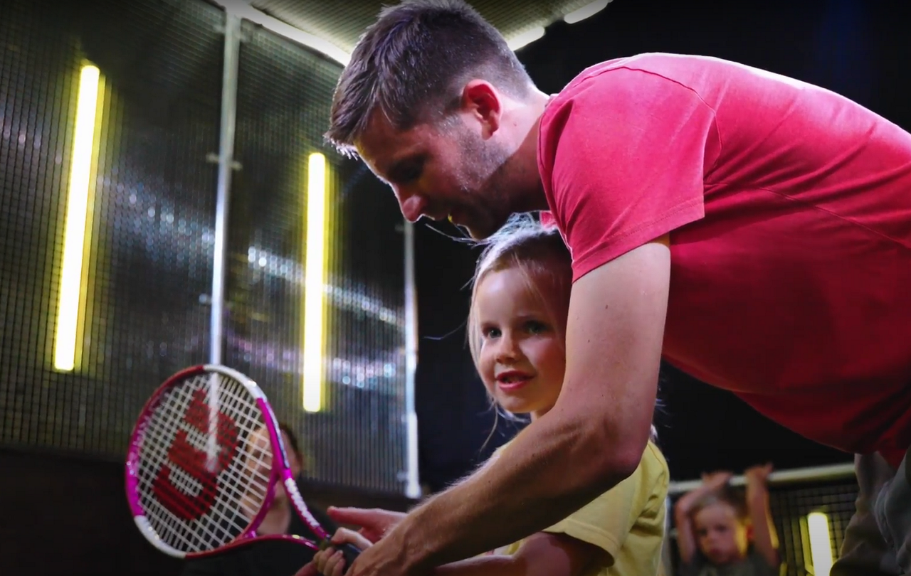 Young girl holding a tennis racket with the help of an adult.