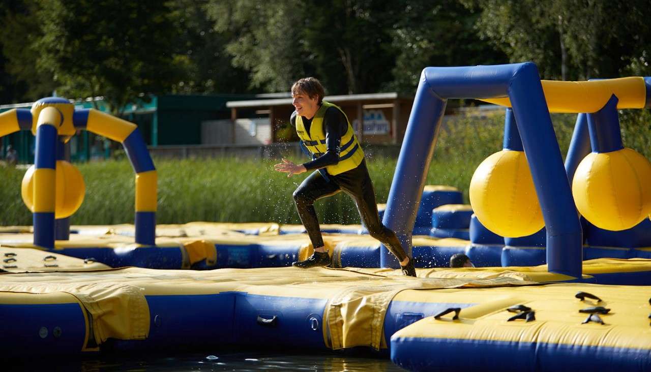Participant runs across a blue-and-yellow inflatable obstacle course, dodging large hanging spheres as water splashes. The course floats on a lake beside reeds, trees, and buildings on a sunny day.