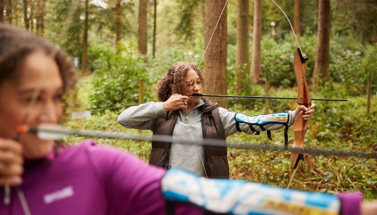 Two archers draw recurve bows, aiming arrows; context: a forest archery range with trees, rope barrier, and armguards, one archer sharply focused mid-frame, another blurred in foreground.