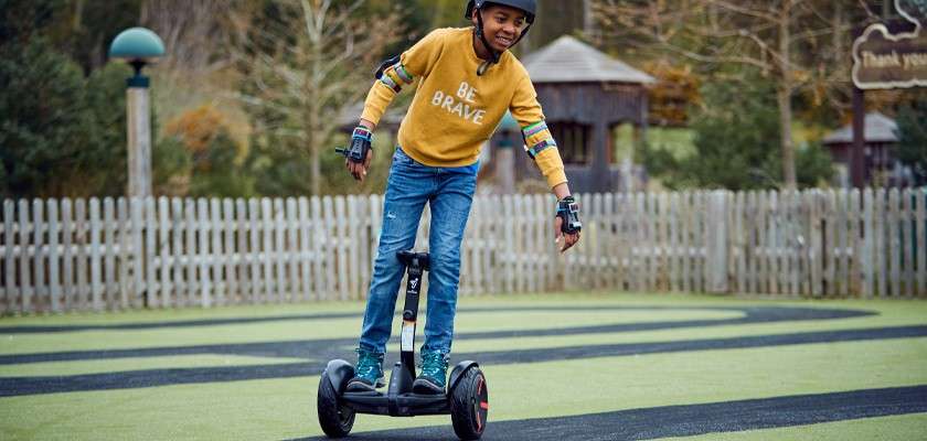 Child rides a two-wheeled self-balancing scooter, leaning forward on a winding track. Park setting with wooden fence and huts. Sweater text: “BE BRAVE.” Distant sign reads: “Thank you.”