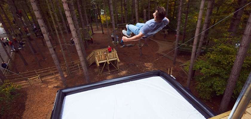 A person flips backward in midair, descending toward a large white airbag. Surrounding context: a forest adventure park with tall trees, wooden platforms, ropes, and scattered onlookers.