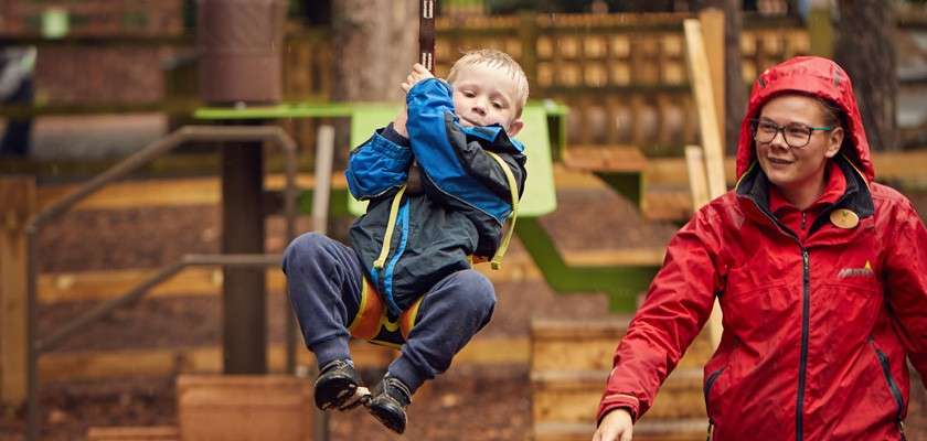 Child rides a zip line, gripping the handle, while an adult in a red rain jacket watches and guides. Wooden platforms and trees surround them in an outdoor playground.