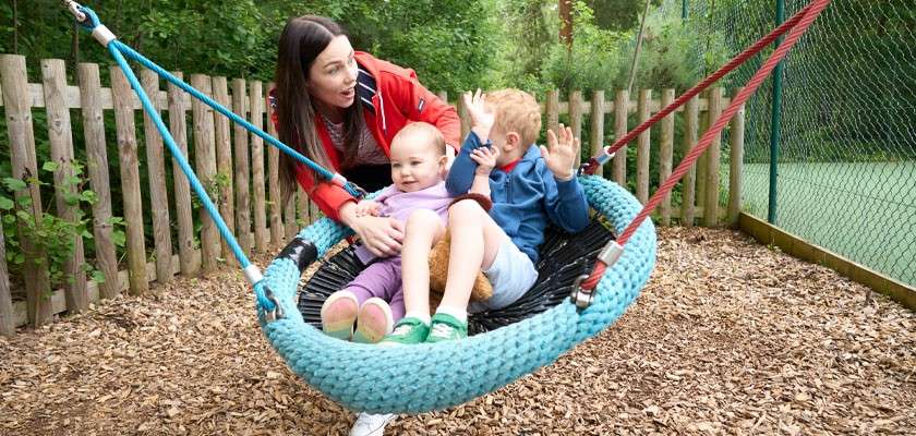 Two children sit on a blue woven basket swing, holding a teddy, while an adult in red pushes; woodchip ground, wooden fence, and trees surround an outdoor playground.