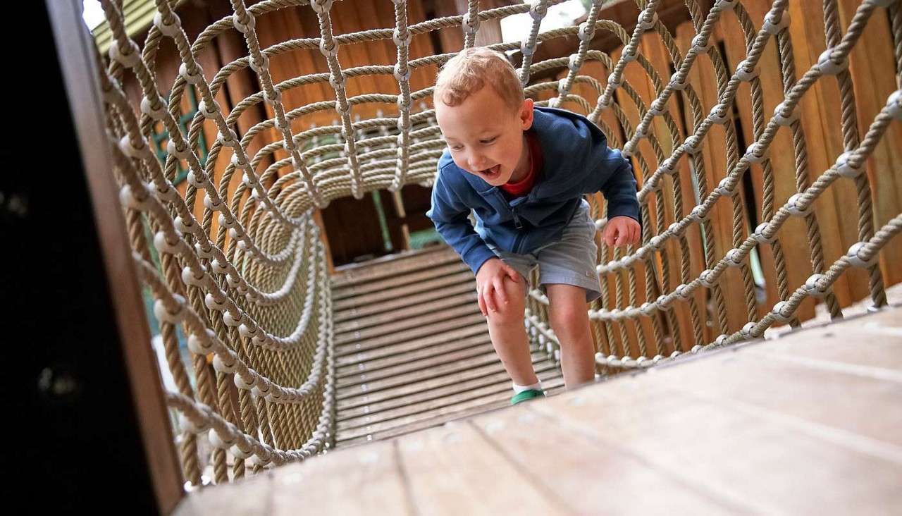 Young boy crossing a rope bridge in an outdoor play area.
