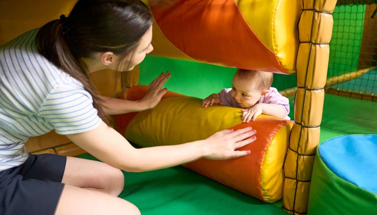 Baby crawls between padded colorful rollers, reaching toward an adult supervising nearby, within an indoor soft play area with green matting and foam structures.