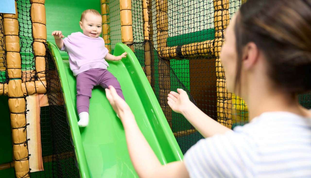 Young girl sliding down a green slide in a soft play area.