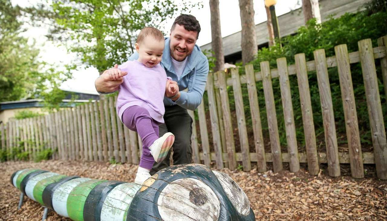 Toddler steps onto a carved caterpillar log while an adult supports their hands. In a playground, surrounded by a wooden fence, trees, and woodchip ground, they practice balance and play.