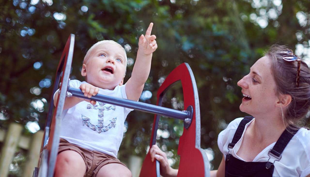 Toddler points upward while gripping a metal bar, riding a red playground rocker; nearby, a smiling woman watches. Sunlit trees blur in background, suggesting an outdoor park setting.