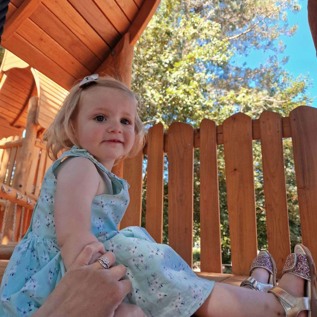 Toddler sits smiling, holding an adult’s hand, on a wooden playground platform. She wears a light blue dress and sparkly shoes. Sunlit trees and a wooden fence surround the structure.