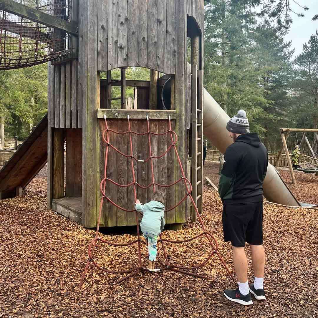 Child climbs a red rope net on a wooden play tower. An adult watches nearby, wearing a beanie labeled "PACK". Surroundings include slide, swings, bark mulch ground, and tall trees.