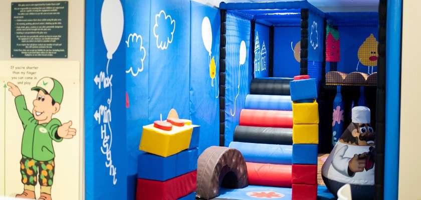 Indoor soft play area climbs padded rainbow steps and navigates foam blocks within a blue, cartoon-decorated room.

Text:
- If you're shorter than my finger you can play.
- Max
- Min Height
- Small policy plaque text unreadable.