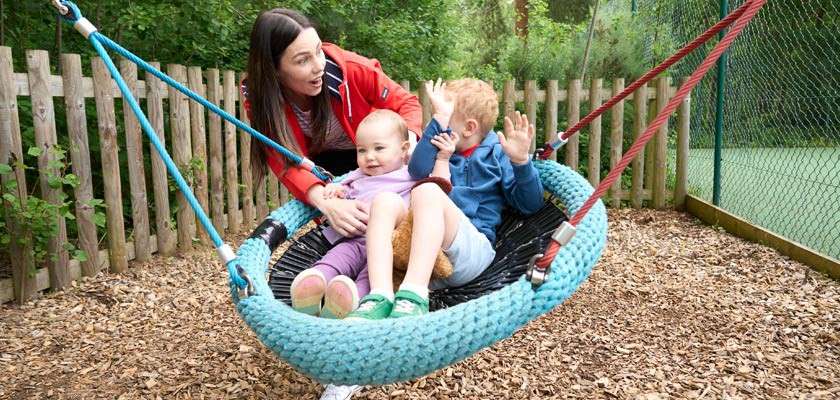 Two small children sit in a large woven basket swing while an adult steadies it, outdoors in a playground with wood-chip ground, wooden picket fence, green netting, and trees.