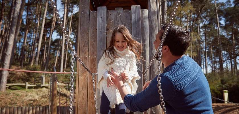 Girl descends a wooden playground platform, smiling, while a man reaches up to steady her. Chain handrails flank the walkway. Sunlit forest and park surroundings provide natural outdoor context.