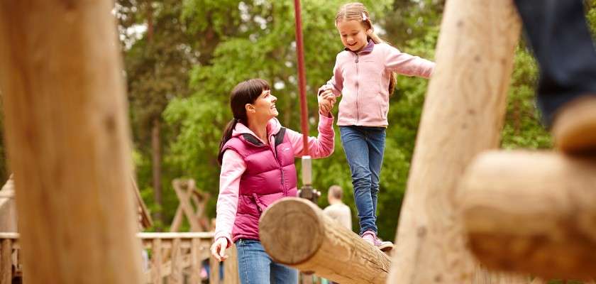Child balances on a round wooden beam, holding an adult’s hand for support, as they navigate a natural playground structure surrounded by trees and other timber equipment.
