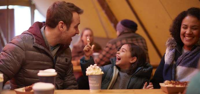 A laughing child raises a spoon, seated between two smiling adults at a wooden table with hot drinks and desserts, inside a warm, tent-like café filled with bundled patrons.