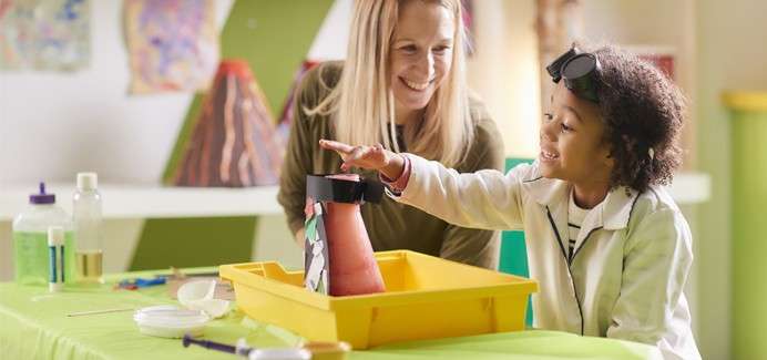 Child in a lab coat reaches toward a model volcano on a tray as an adult supervises in a classroom, surrounded by beakers, droppers, goggles, and colorful art projects.