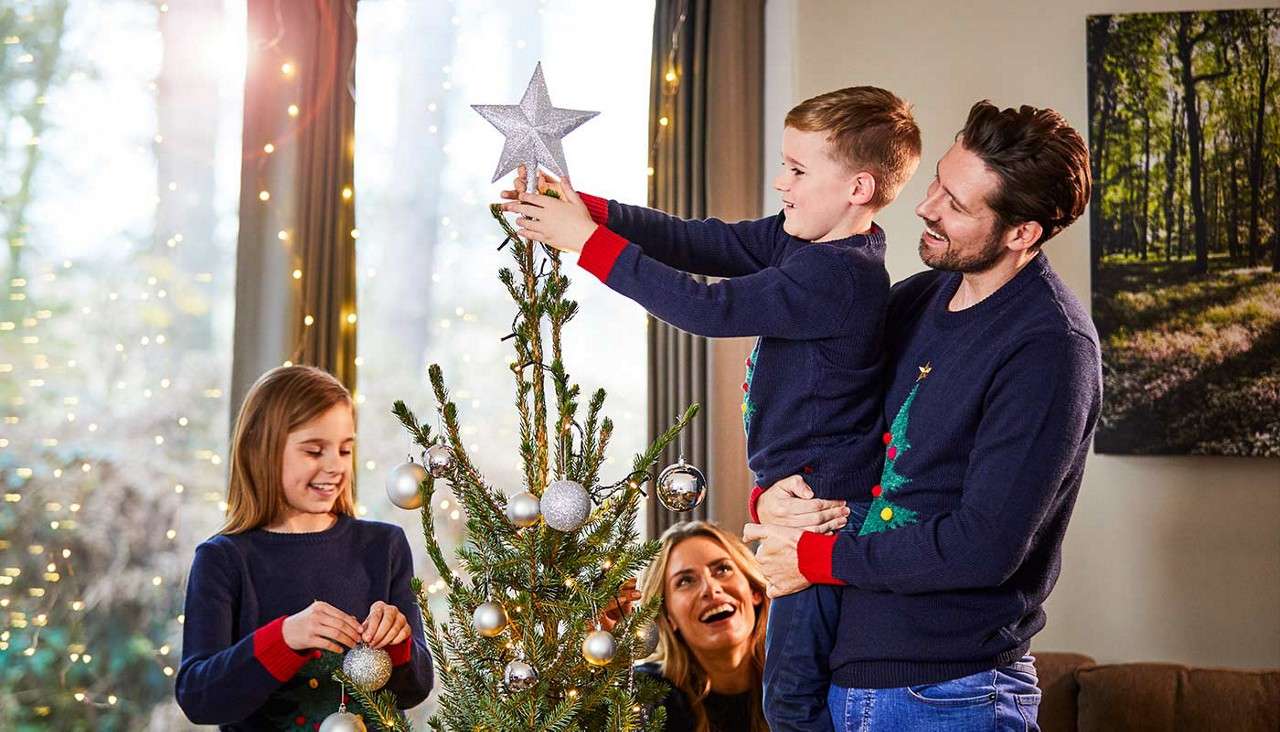Boy places a star topper on a Christmas tree, supported by a man; a girl hangs ornaments while a woman watches, in a cozy living room with fairy lights and daylighted windows.