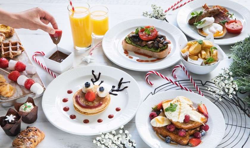 Breakfast dishes sit arranged; a hand dips a strawberry into chocolate. Plates show pancakes, toast with mushrooms, eggs with bacon, French toast, juices, potatoes, waffles, berries on a white table.