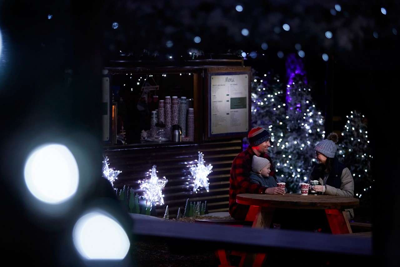 A couple sat on a bench in the dark with hot drinks surrounded by festive lights.