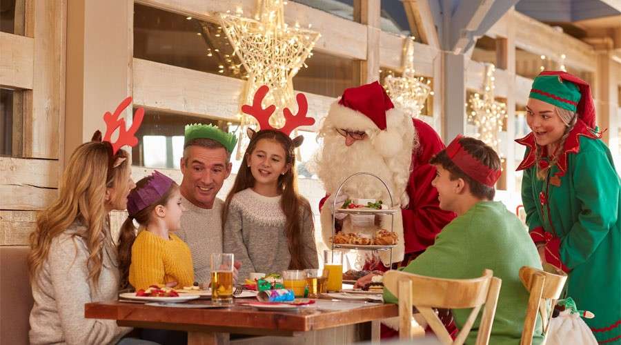 Santa and an elf present a tiered tray of treats to a smiling family. They dine at a wooden table, wearing festive headbands, inside a warmly lit, holiday-decorated restaurant.