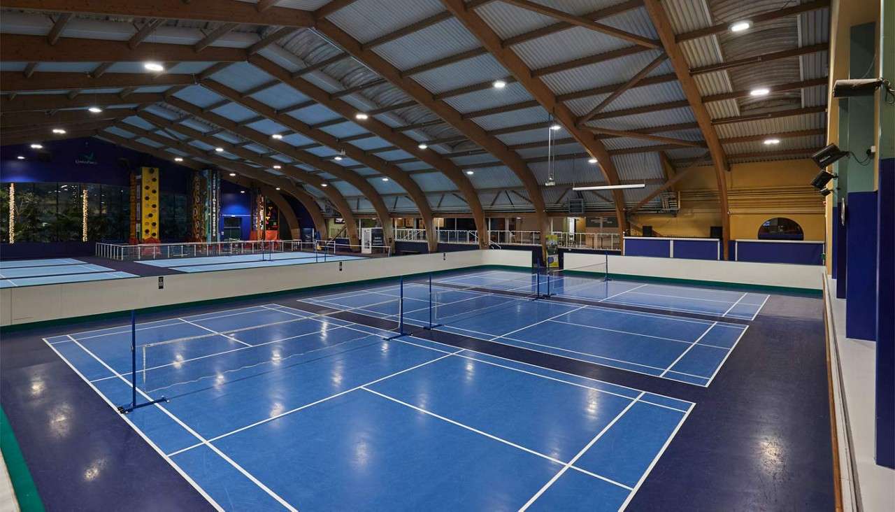 Badminton courts sit empty with nets strung across blue floors, under arched wooden beams. Nearby, raised walkways, seating, and colorful climbing walls occupy the spacious indoor sports facility.