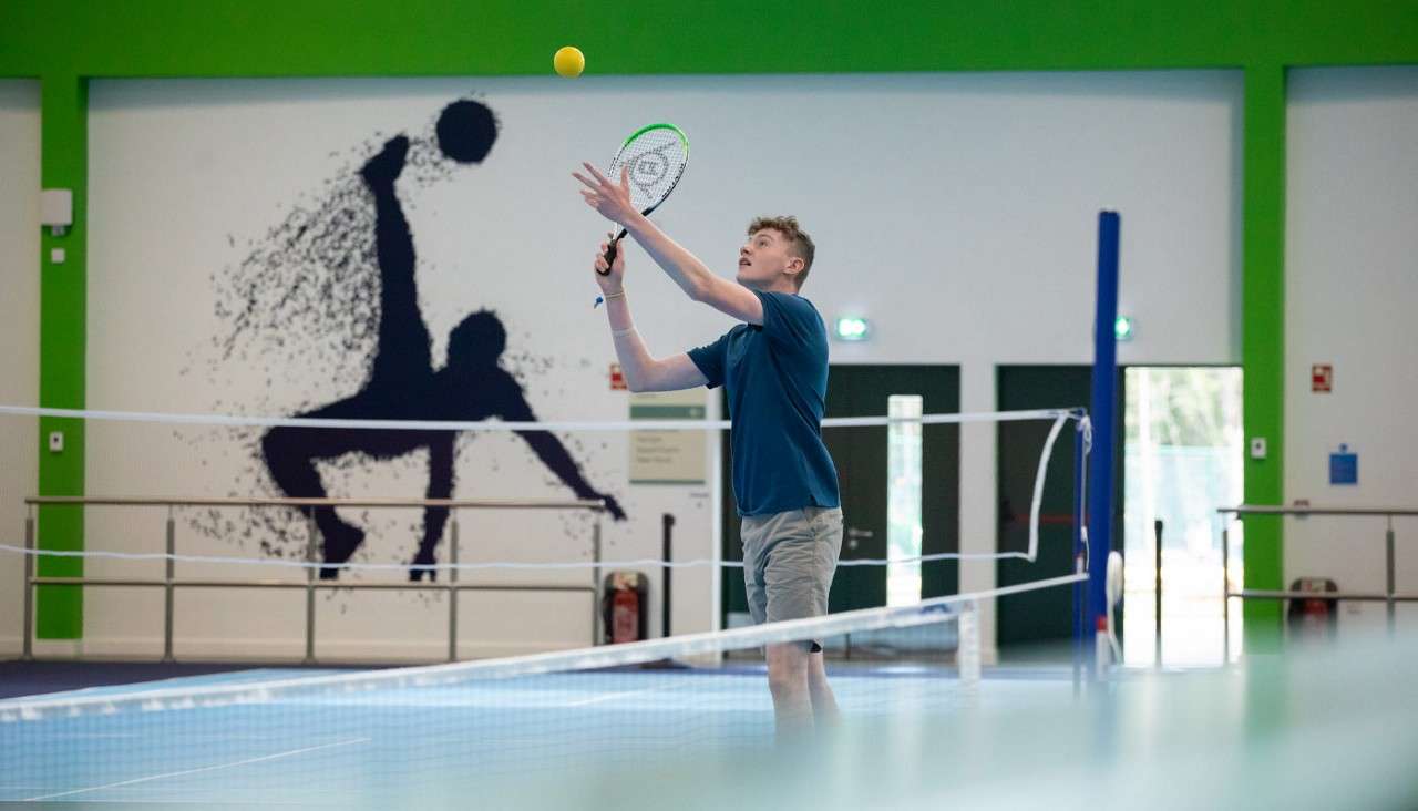 Player tosses a yellow tennis ball to serve, racket raised, on an indoor court with low net, green-and-white walls, doors, and a large athlete silhouette mural.
