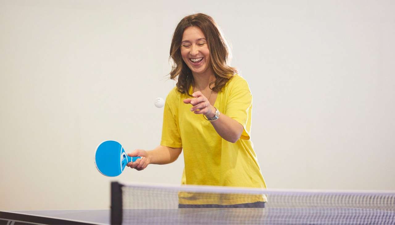 Smiling person hits a ping-pong ball with a blue paddle, mid-serve, standing behind a table-tennis net in a bright indoor room with plain walls.
