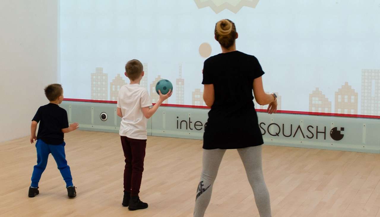 Adult and two children stand and play with a ball, facing an interactive projection on a squash court with cityscape graphics. Text on the glass wall reads: inte SQUASH.