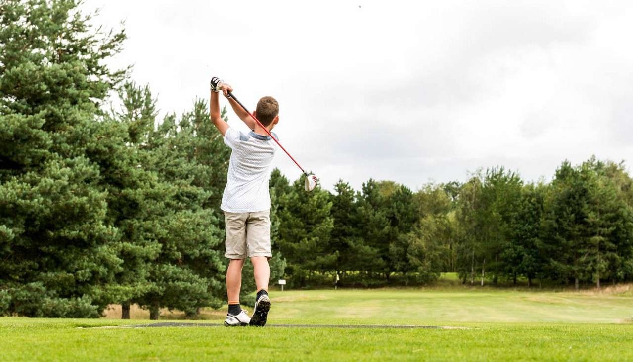 A youth golfer swings a driver through his follow-through, teeing off on a manicured fairway, bordered by dense pine trees beneath an overcast sky.