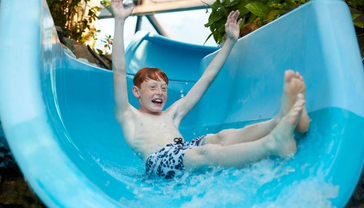 Child raises arms while sliding down a blue water slide, splashing water, smiling; indoor waterpark setting with plants and structural beams visible around the slide.