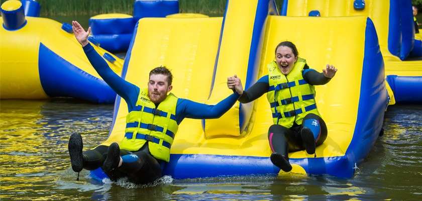 Two adults in yellow life vests hold hands while sliding down a bright inflatable water slide, splashing into a lake, surrounded by other blue-yellow floating obstacles.