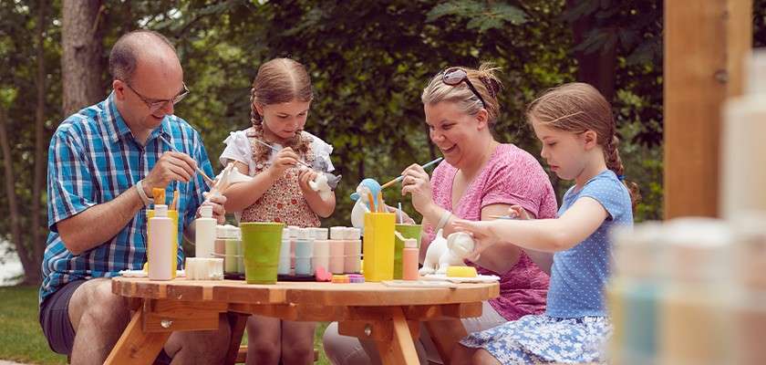 A family sat at a wooden picnic table outside painting pottery together