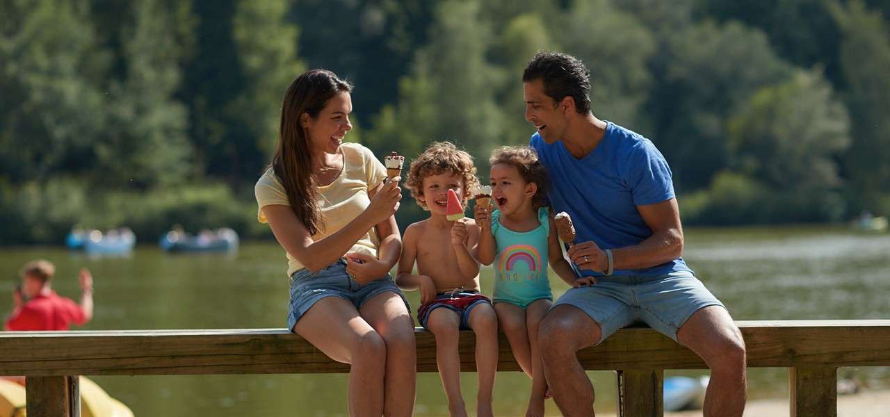 Family of four sits on a wooden railing, eating ice creams and popsicles, smiling together. Background shows a calm lake with paddle boats and trees. Text visible: "HELLO" on the child’s swimsuit.