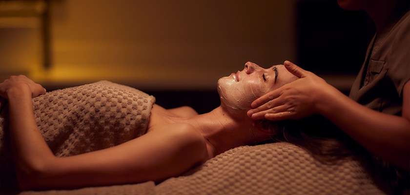 Person receives a facial treatment; therapist massages cheeks while a mask covers the face, on a textured blanket in a dim, warm spa room.
