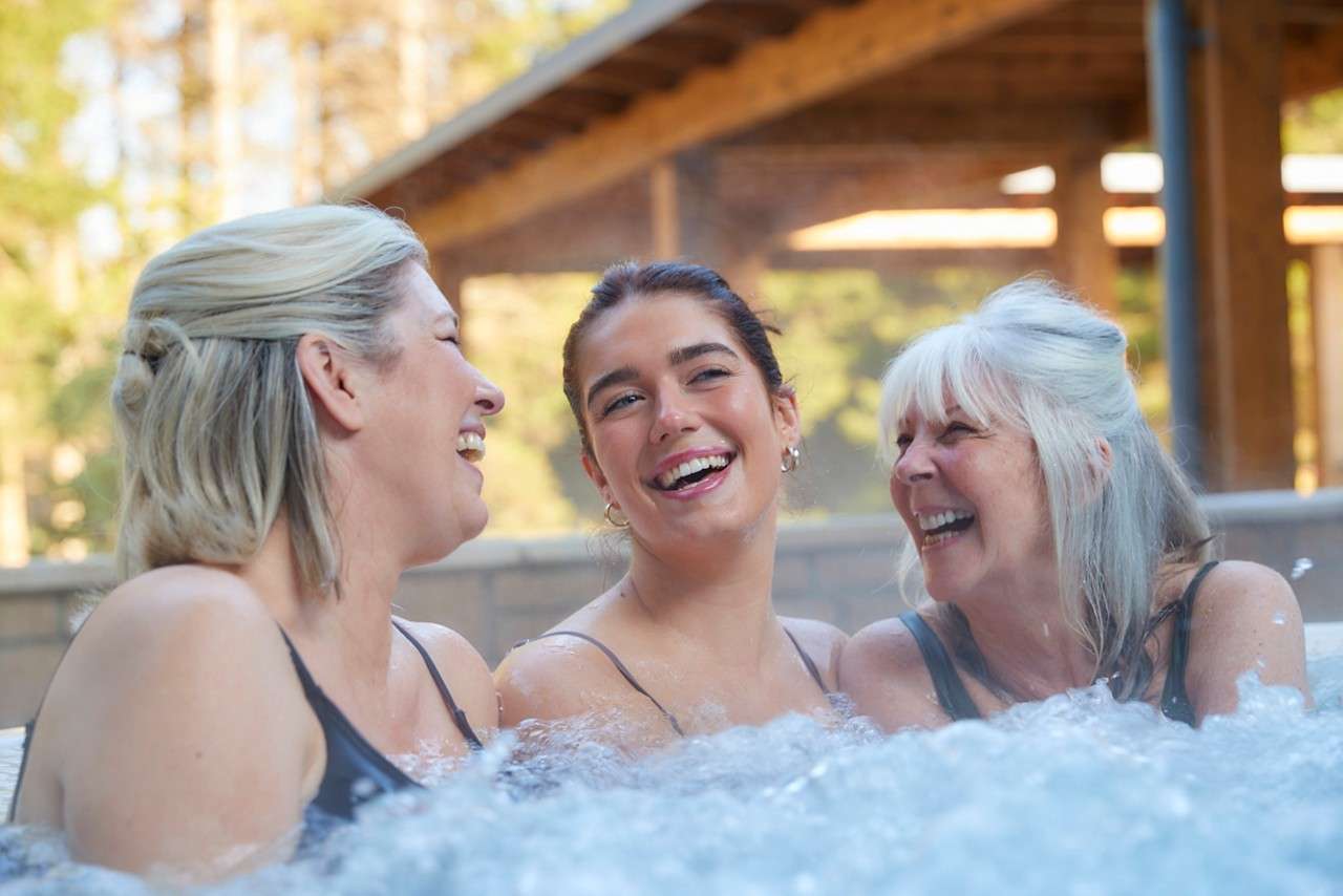 Three people relax and laugh, immersed shoulder-deep in a bubbling hot tub, outdoors at a spa beneath a wooden pergola, with sunlit trees in the background.