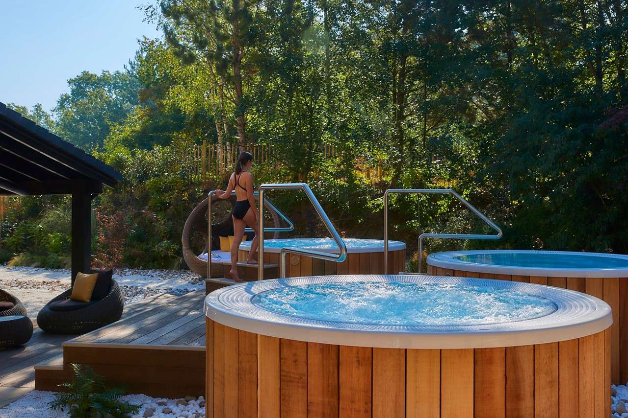 Person in a swimsuit steps into a bubbling circular hot tub, holding a metal handrail, on a wooden outdoor spa deck with multiple tubs, lounge chairs, and surrounding trees.