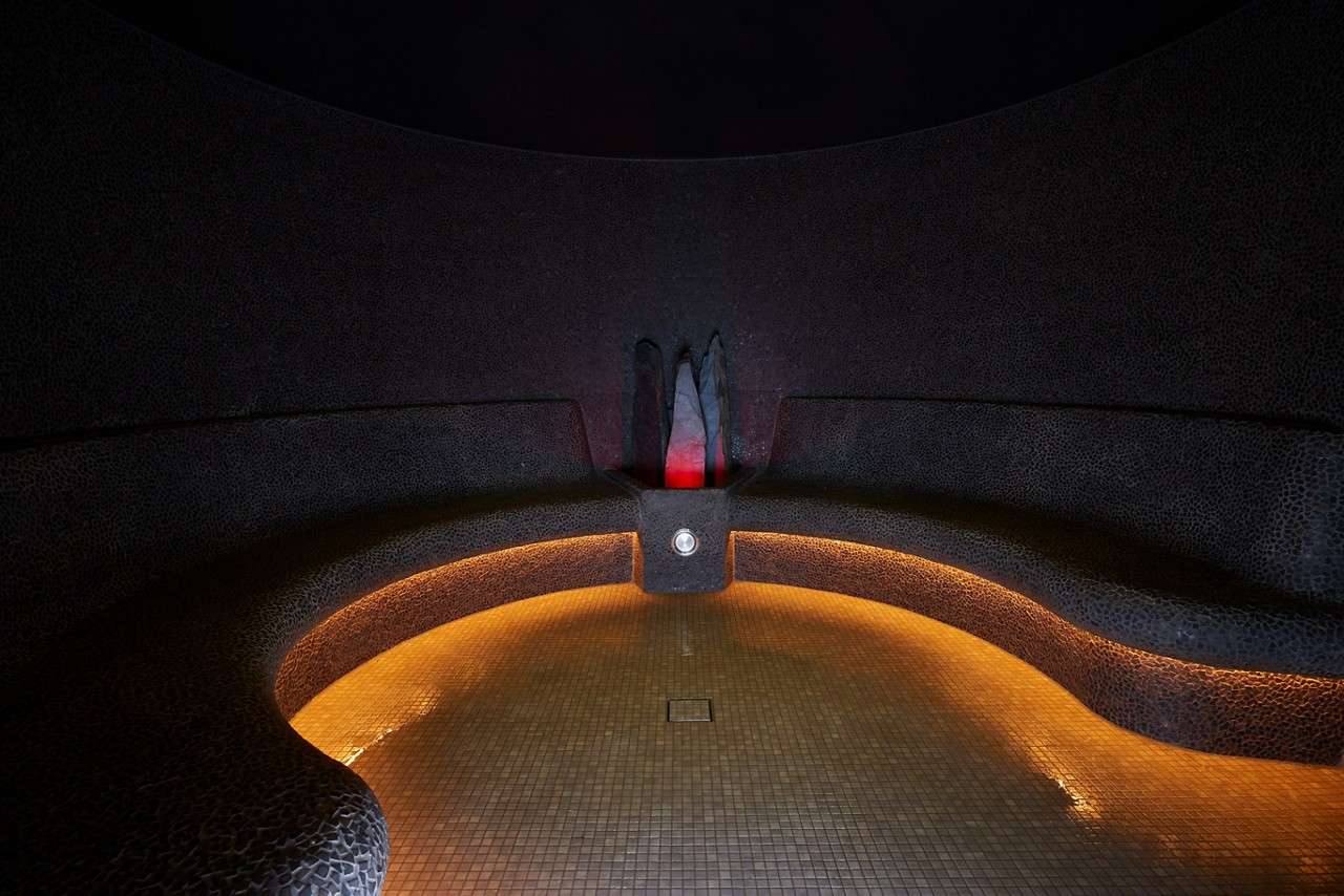 Curved tiled bench glows with amber underlighting, encircling a central steam outlet beneath stone fins, while faint red light rises; set in a dim, mosaic-tiled spa steam room.