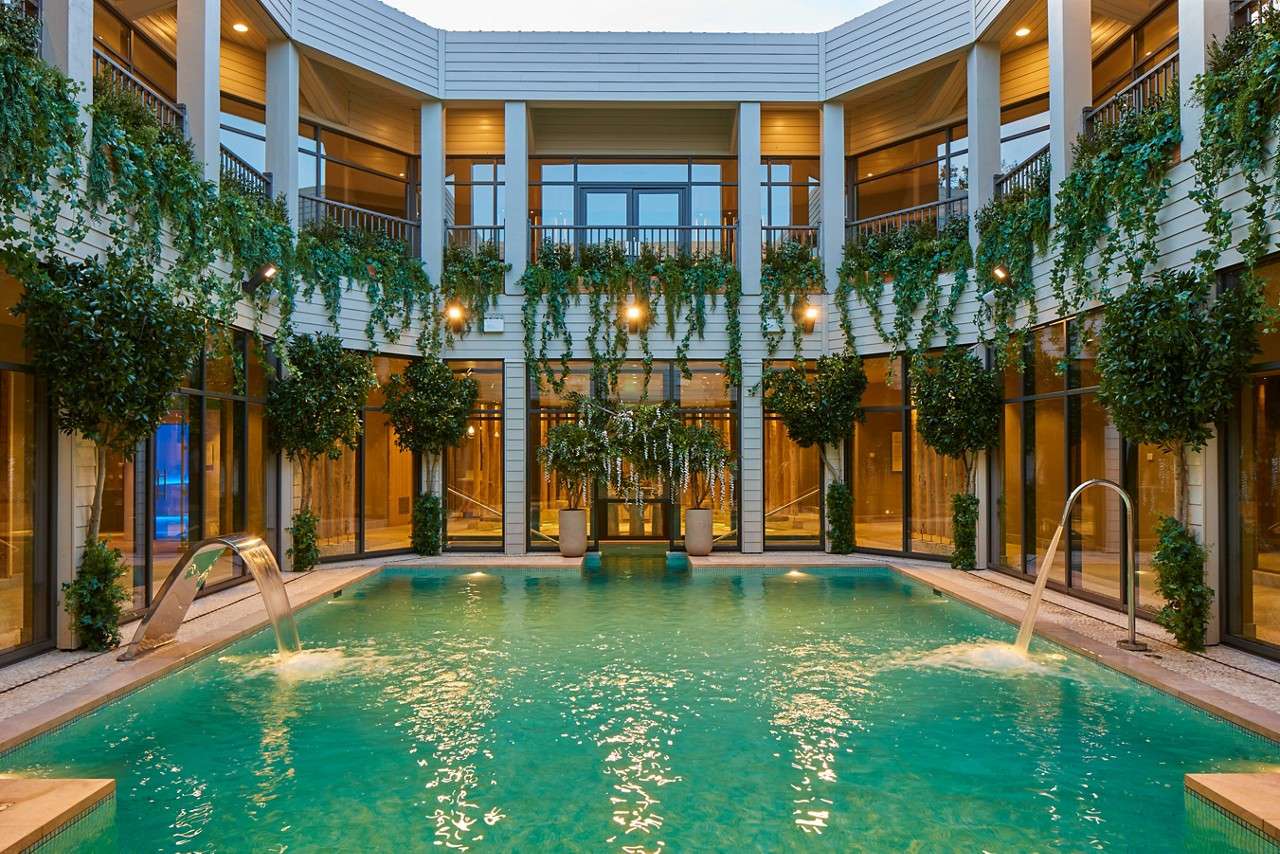 Swimming pool bubbles and flows from metal spouts, in a circular two-story courtyard. Glass walls, balconies, hanging vines, and potted trees surround the water under warm ambient lighting.