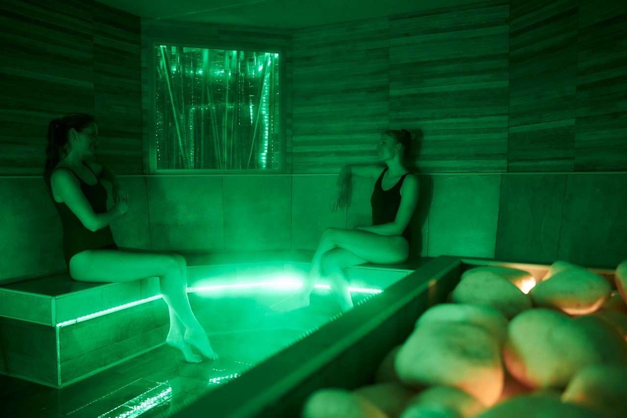 Two people sit and talk, feet near illuminated water, in a green-lit spa steam room with tiled benches and a tray of hot stones in the foreground.