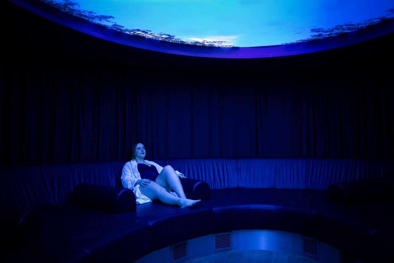 Person reclines on a curved leather bench under blue ambient light in a circular dark room with black curtains and a round illuminated ceiling feature, suggesting a spa-like relaxation space.